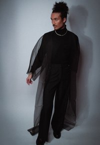 Veste kimono noire en tissu transparent par-dessus un col roulé noir et un pantalon. Manches longues, coupe ample, détails de boutons subtils. Texture lisse sur toute la surface.