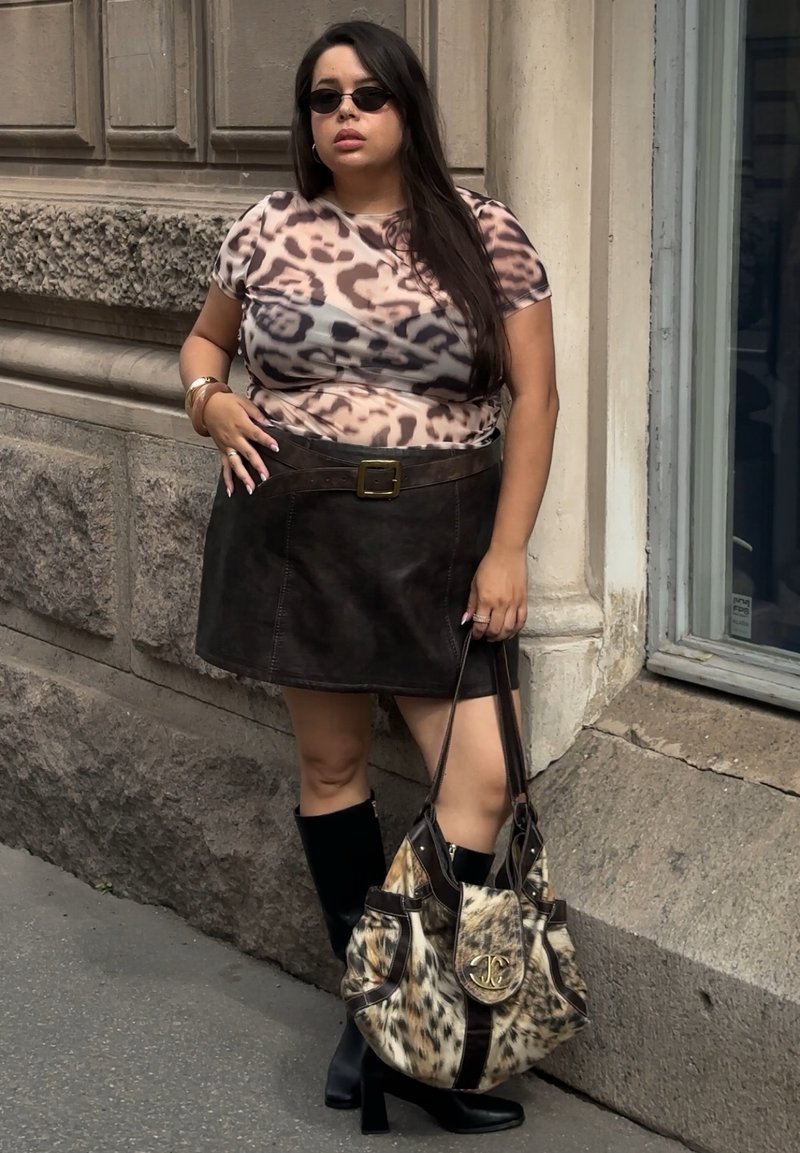 Femme aux longs cheveux foncés portant des lunettes de soleil, un haut à imprimé léopard, une jupe noire et des bottes, tenant un grand sac à main à motif animalier dans une rue de la ville.