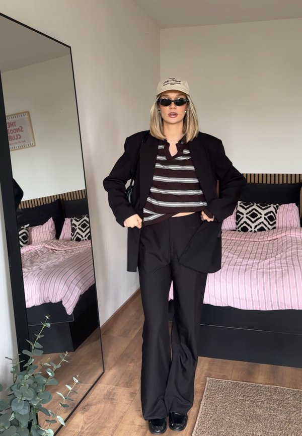 Mujer con blazer negro, camisa de rayas, pantalones negros, gafas de sol y gorra beige, de pie en un dormitorio con una cama y un espejo de cuerpo entero.