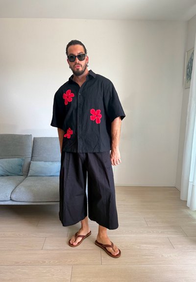 Hombre con pantalones negros de pierna ancha, una camisa negra con flores rojas, gafas de sol y chanclas marrones de pie en una sala de estar luminosa.