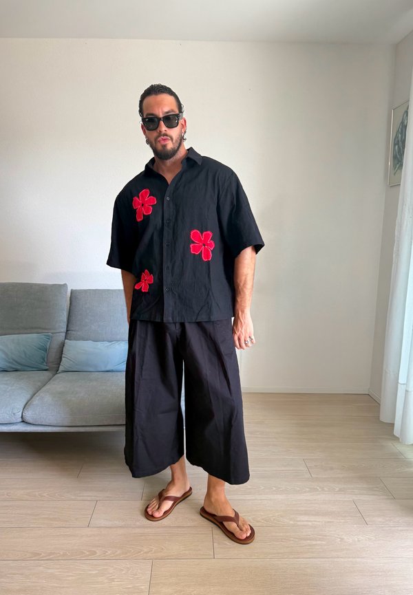 Mand iført sorte vidde bukser, en sort skjorte med røde blomster, solbriller og brune flip-flops, der står i en lys stue.
