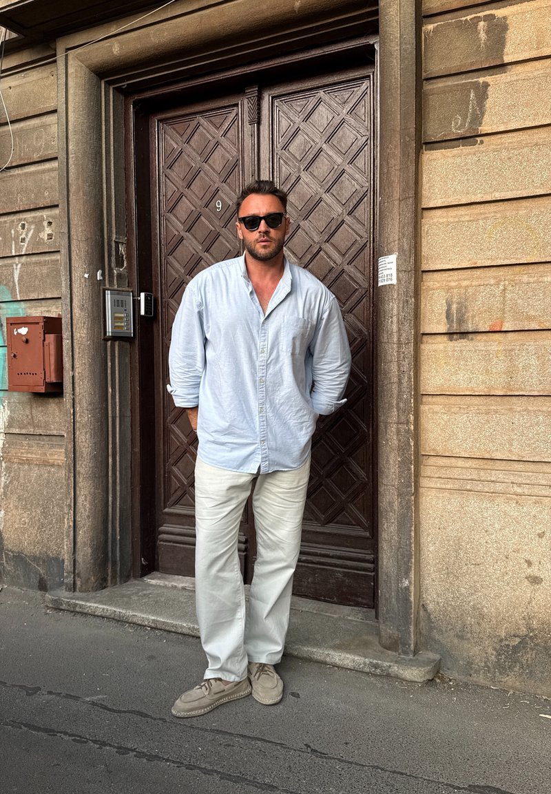 Homme portant des lunettes de soleil, une chemise bleu clair et un pantalon blanc, debout les mains dans les poches devant une grande porte en bois avec des motifs en losange.