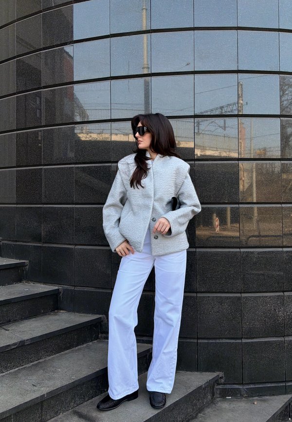 Woman with long dark hair wearing black sunglasses, a grey coat, white trousers, and black shoes standing on grey steps against a dark tiled wall.