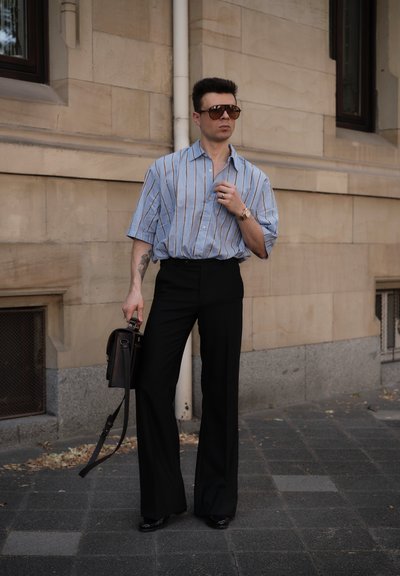Camisa de manga corta a rayas en azul claro; pantalones anchos negros; gafas de sol; sosteniendo un bolso marrón oscuro. Entorno urbano con muros de piedra.