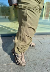 Person mit olivgrünen Hosen und beigen Nike-Sneakern, der auf einem grauen gefliesten Bürgersteig im Sonnenlicht steht.