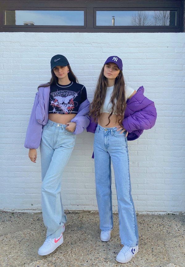 Dos mujeres están de pie frente a una pared de ladrillos blancos. Una lleva una chaqueta acolchada púrpura, un top corto negro y jeans de mezclilla claros; la otra viste un top corto blanco, jeans de tiro alto y una gorra púrpura. Ambas llevan zapatillas.