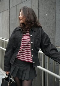 Femme aux cheveux bouclés portant une veste en jean noire, un chemisier rayé bordeaux, une jupe plissée, tenant un sac à main noir près d'un mur en béton gris.