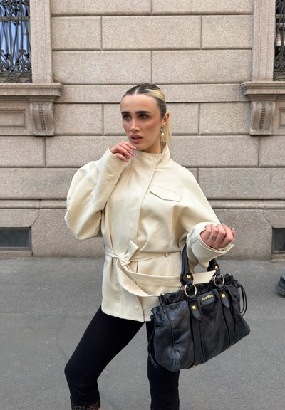 Mujer con el pelo peinado hacia atrás que lleva una chaqueta beige con cinturón y pantalones negros sostiene un bolso negro de Miu Miu, de pie junto a la pared de un edificio de piedra.