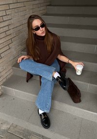 Barna bordázott garbó, világoskék bővülő farmernadrág, fekete arany díszítésű mokaszin, fehér zokni, kávéscsészét tartva, szürke lépcsőkön ülve.