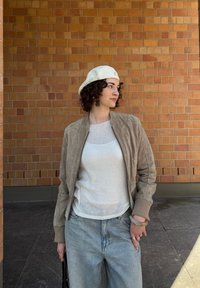 Jaqueta bomber de camurça bege, blusa de malha branca, jeans de ganga azul claro. Os acessórios incluem um boné branco e joalharia. Fundo texturizado de tijolo.