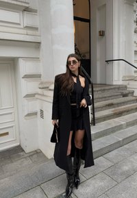 Černý trenčkot přes černý top a minisukni, doplněný lesklými černými kozačkami po kolena. Doplňky zahrnují stylové sluneční brýle a malou černou kabelku.