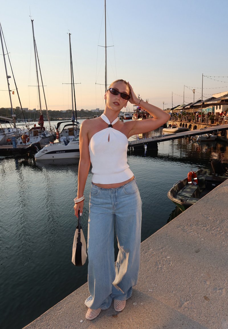 Femme portant un haut à col montant blanc, un jean à jambes évasées et des lunettes de soleil, debout près de la marina avec des voiliers et des restaurants en bord de mer au coucher du soleil.