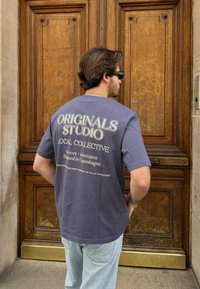 Γκρίζο T-shirt με άνετη εφαρμογή, με λευκό κείμενο στο πίσω μέρος που γράφει "ORIGINALS STUDIO LOCAL COLLECTIVE." Κλασικός σχεδιασμός με στρογγυλή λαιμόκοψη.