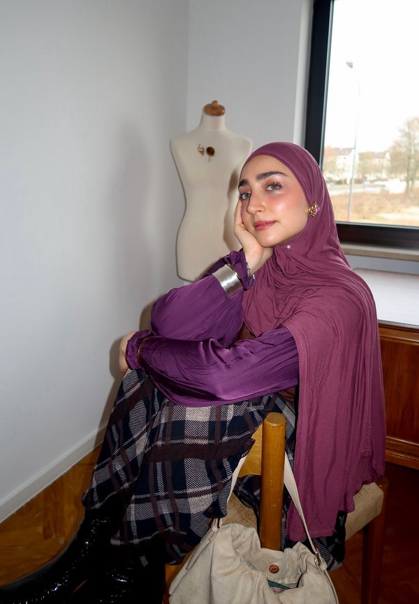 Vrouw met een paarse hijab en blouse zittend op een stoel, haar hoofd steunend op haar hand, met een geruite rok en een paspop op de achtergrond.