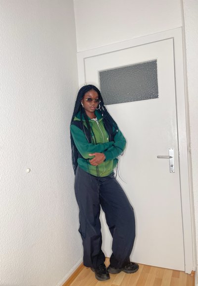 Chaqueta de cremallera verde y negra, pantalones cargo negros y zapatillas negras. Cabello largo trenzado, de pie frente a una puerta blanca con panel de vidrio.