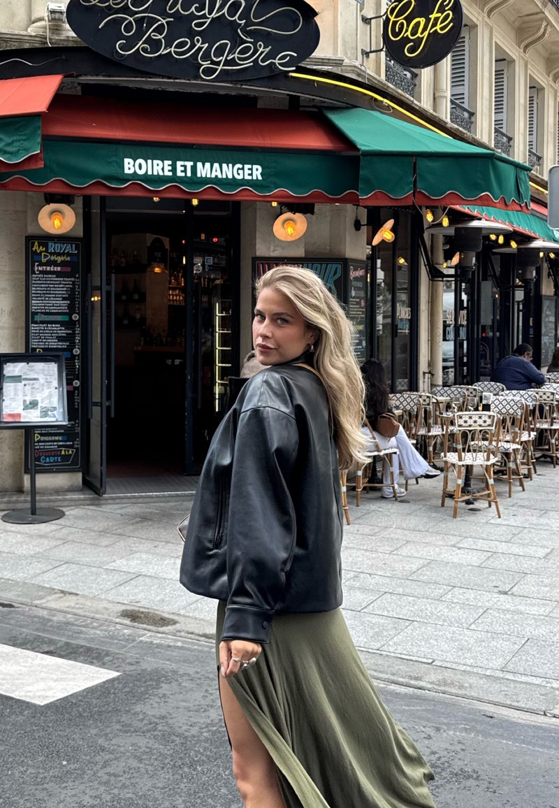 Schwarze Bomberjacke aus Leder, grüner Maxirock mit Oberschenkelschlitz, lange blonde Haare, steht vor einem Café mit Außensitzplatz.