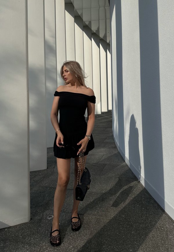 Zwart off-the-shoulder jurk met een uitlopende zoom, gecombineerd met zwarte netten schoenen en een gewatteerde handtas, tegen een achtergrond van witte architectonische zuilen.