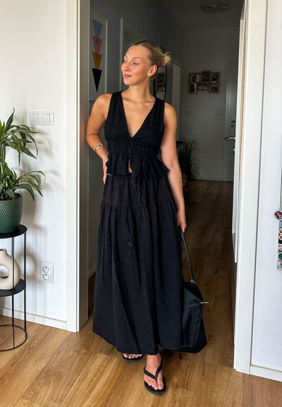Vestido maxi negro en capas con escote en V profundo, cintura con volantes y un corte holgado. La modelo sostiene un bolso negro y lleva chanclas.