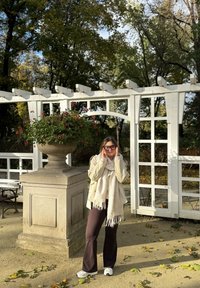 Vaaleanbeige takki ja kermaisenvärinen huivi, joita käyttää henkilö harmaan kivisen kukkalaatikon vieressä, jossa on punaisia kukkia, taustalla puistossa puita.