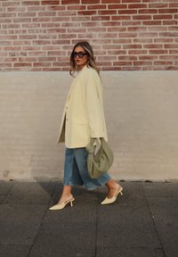 Γυναίκα που περπατά δίπλα σε τοίχο από τούβλα, φορώντας φαρδιά παλτό σε παλ κίτρινο, κομμένα τζιν, κίτρινες σανδάλια με τακούνι, γυαλιά ηλίου και κρατώντας μια πράσινη τσάντα.