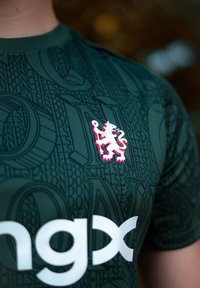 Zaļš sporta T-krekls ar teksturētu rakstu, balts logo un izšūts lauvas emblēma sarkanā un baltā krāsā, novietots kreisajā krūtī.