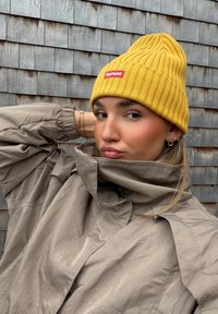 Bonnet jaune à côtes avec une étiquette rouge, porté avec une veste beige clair froissée. L'arrière-plan présente des bardeaux en bois.