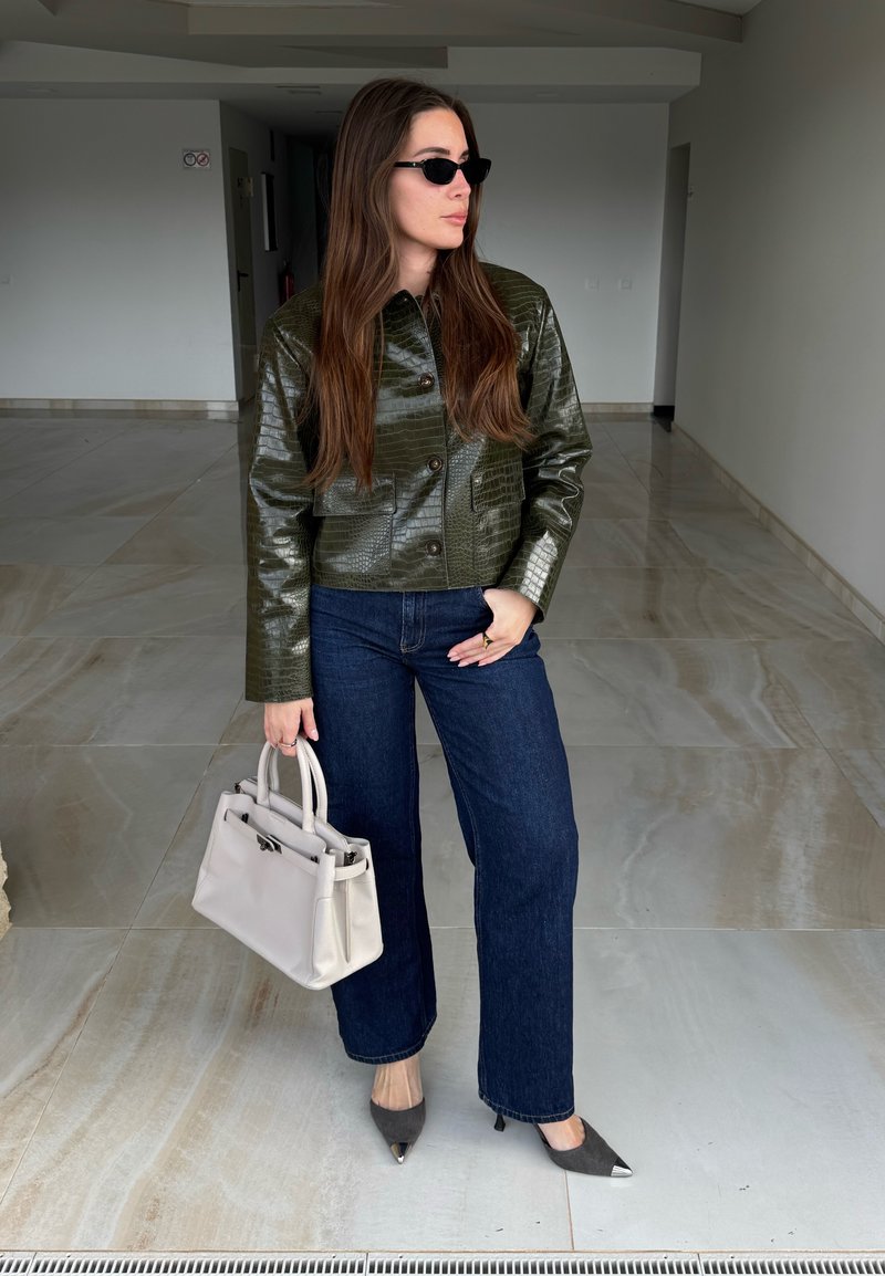 Femme aux longs cheveux bruns portant des lunettes de soleil foncées, une veste verte texturée, un jean bleu, des talons noirs pointus, tenant un sac à main blanc.