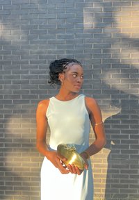 Svetlomodré šatôčky bez rukávov s hladkou textúrou; drží lesklú zlatú kabelku s kvetinovým akcentom; stojí vedľa šedej tehlovej steny.