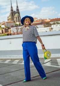 Mulher a caminhar ao ar livre, vestida com um chapéu azul, uma camisa às riscas, calças azuis, sapatos verdes, óculos de sol amarelos, um colar colorido e a segurar uma bolsa em forma de limão.
