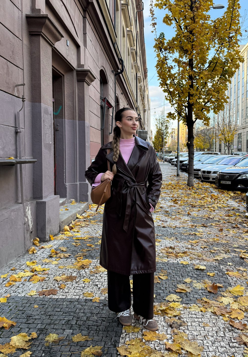 Brown leather trench coat with a belted waist, layered over a pink jumper. Wide black trousers and brown trainers. Autumn leaves on the ground.