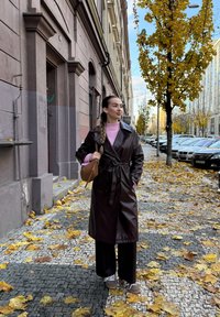Brown leather trench coat with a belted waist, layered over a pink jumper. Wide black trousers and brown trainers. Autumn leaves on the ground.