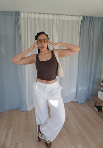 Mujer con trenza en el cabello que lleva gafas de sol naranjas, top sin mangas marrón, pantalones blancos y zapatillas, posando en interiores con las manos cerca de la cabeza.