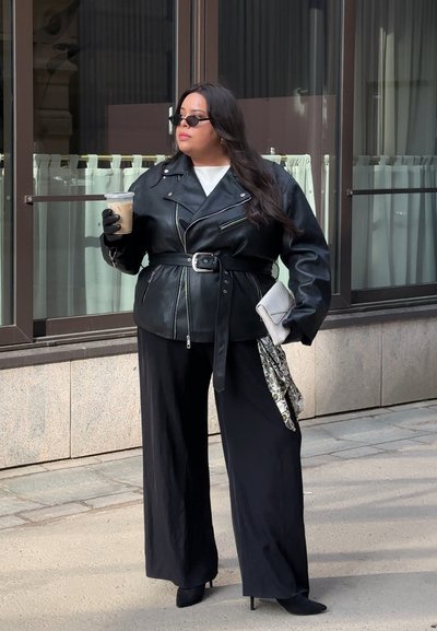 Mujer con chaqueta de cuero negra, pantalones de pierna ancha y guantes sosteniendo café helado y un bolso de mano, de pie en la acera junto a las ventanas de un edificio.