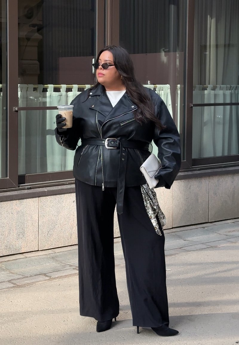 Femme en veste en cuir noire, pantalon à jambes larges et gants, tenant un café glacé et une pochette, debout sur le trottoir près des fenêtres d'un bâtiment.