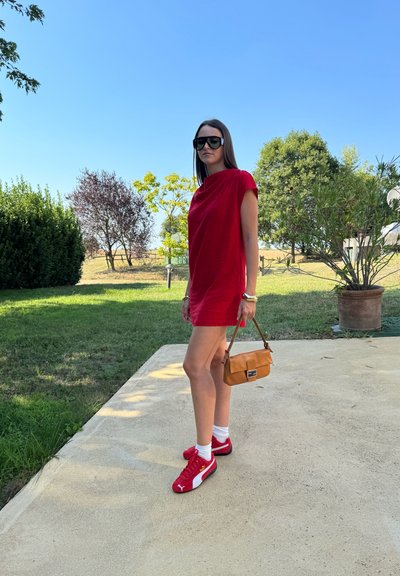Mujer con un vestido rojo corto, zapatillas rojas, calcetines blancos y gafas de sol negras, sosteniendo un bolso marrón, de pie afuera en un día soleado.