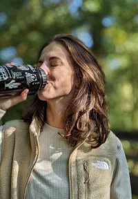 Schwarz isolierte Reisetasse mit weißen Grafiken, nah am Gesicht gehalten. Beigefarbene Fleece-Weste mit silbernem Logo und strukturiertem Pullover.