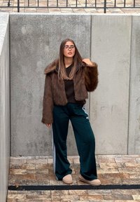 Brun kort jacka i fuskpäls, svart topp, gröna mjukisbyxor med vita ränder och beige tofflor, mot en bakgrund av texturerad betong.