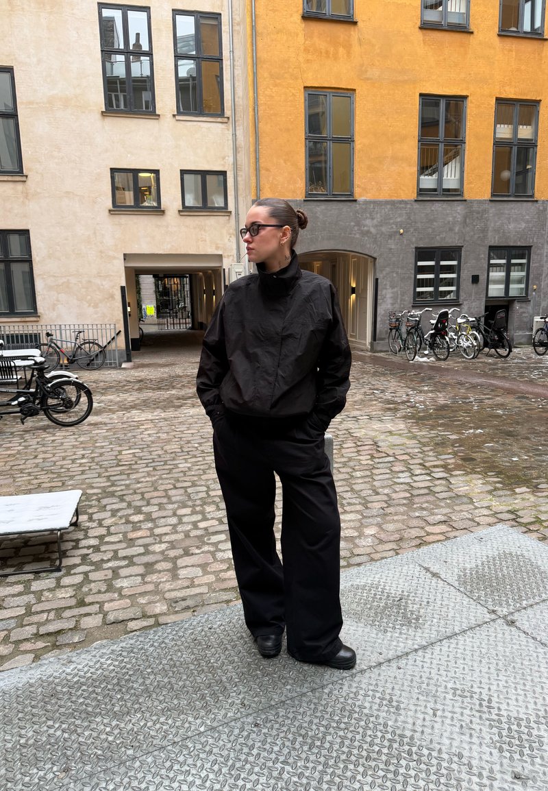 Personne en veste noire et pantalon large se tient sur une rue pavée, les mains dans les poches, près de vélos et de bâtiments urbains à plusieurs étages.