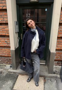 Temno modri plašč iz volnene mešanice z ovratnikom, bela pletena majica, sive kavbojke širokih nog in črni usnjeni gležnarji. V roki črna torba iz blaga.