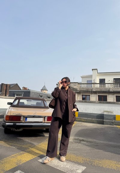 Mujer con ropa oscura y gafas de sol junto a un Mercedes 380 SL beige de estilo vintage en un aparcamiento urbano soleado.