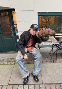 Homme portant une casquette, une veste noire et un jean délavé, assis sur une chaise à l'extérieur d'un café, à côté d'une table avec des fleurs violettes en pot et un sac noir.