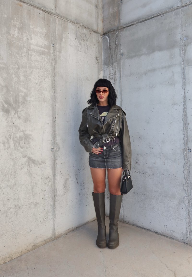 Veste de motard en cuir olive avec quincaillerie argentée, t-shirt graphique noir, mini jupe en denim, bottes vert olive à hauteur de genou et sac à main noir.
