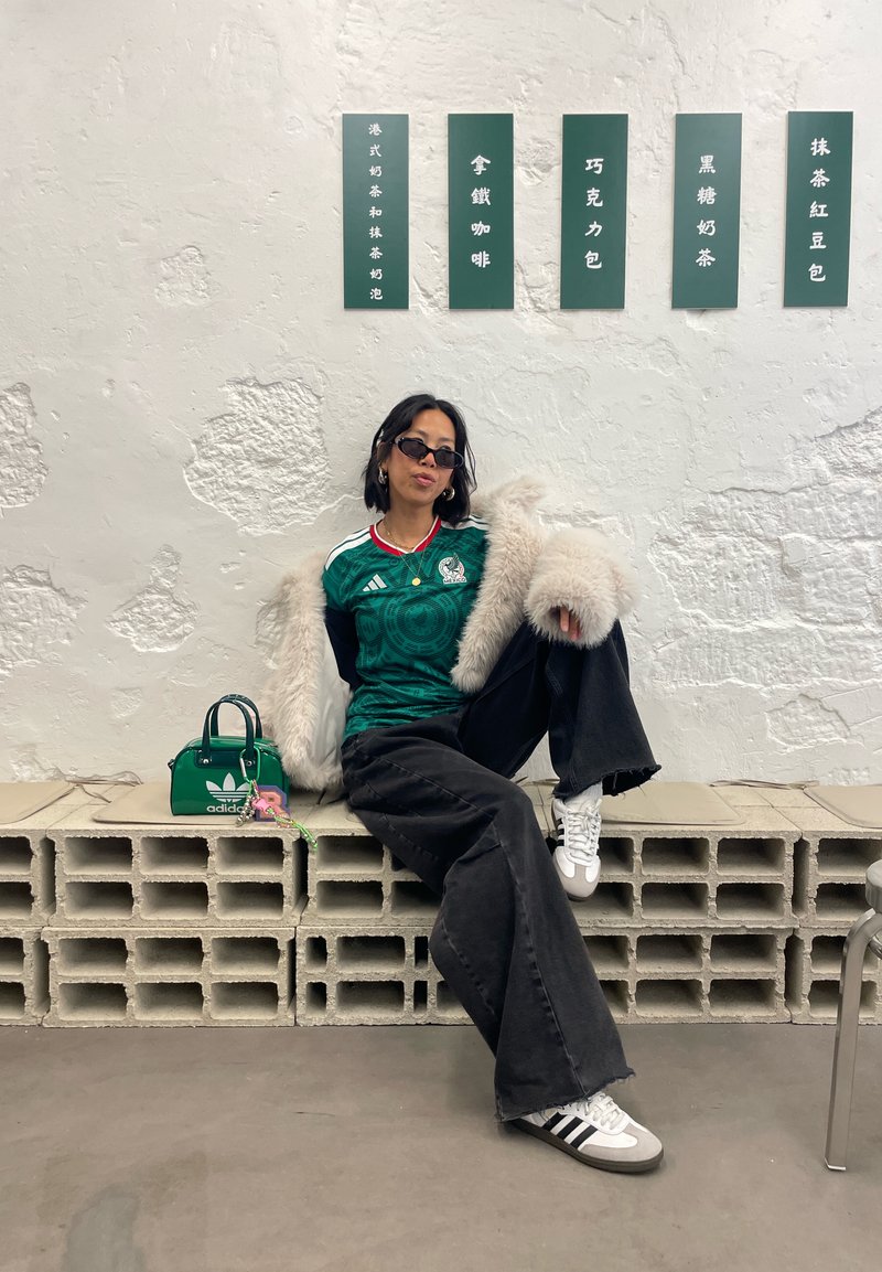 Jeune femme portant des lunettes de soleil, un maillot de sport vert, une veste en fourrure et un pantalon noir, assise sur des blocs de béton à côté d'un sac Adidas vert.