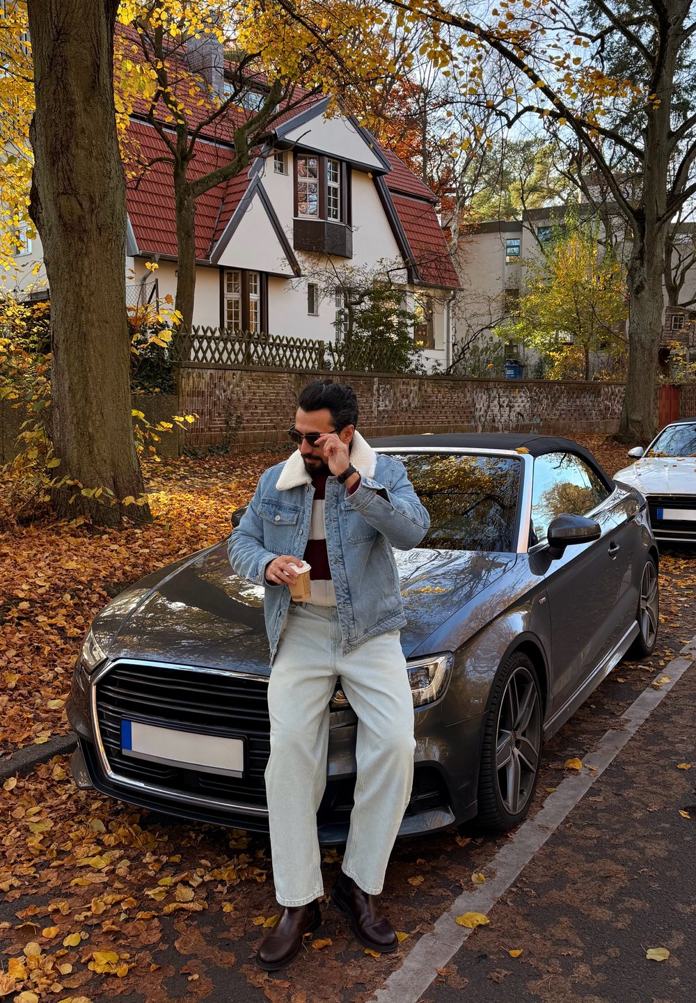 Mann i denimjakke og solbriller som holder kaffekopp, lent mot grå kabrioletparkert på en gate dekket av høstløv.