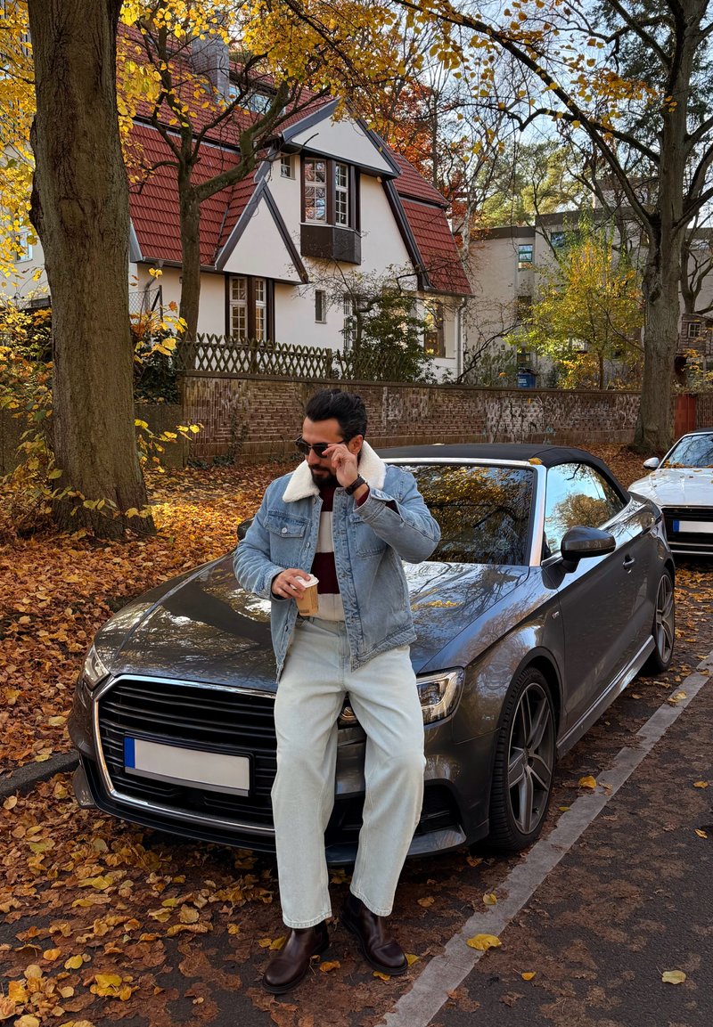 Man in spijkerjack en zonnebril die koffie vasthoudt, leunend op een grijze cabriolet geparkeerd op een met herfstbladeren bedekte straat.