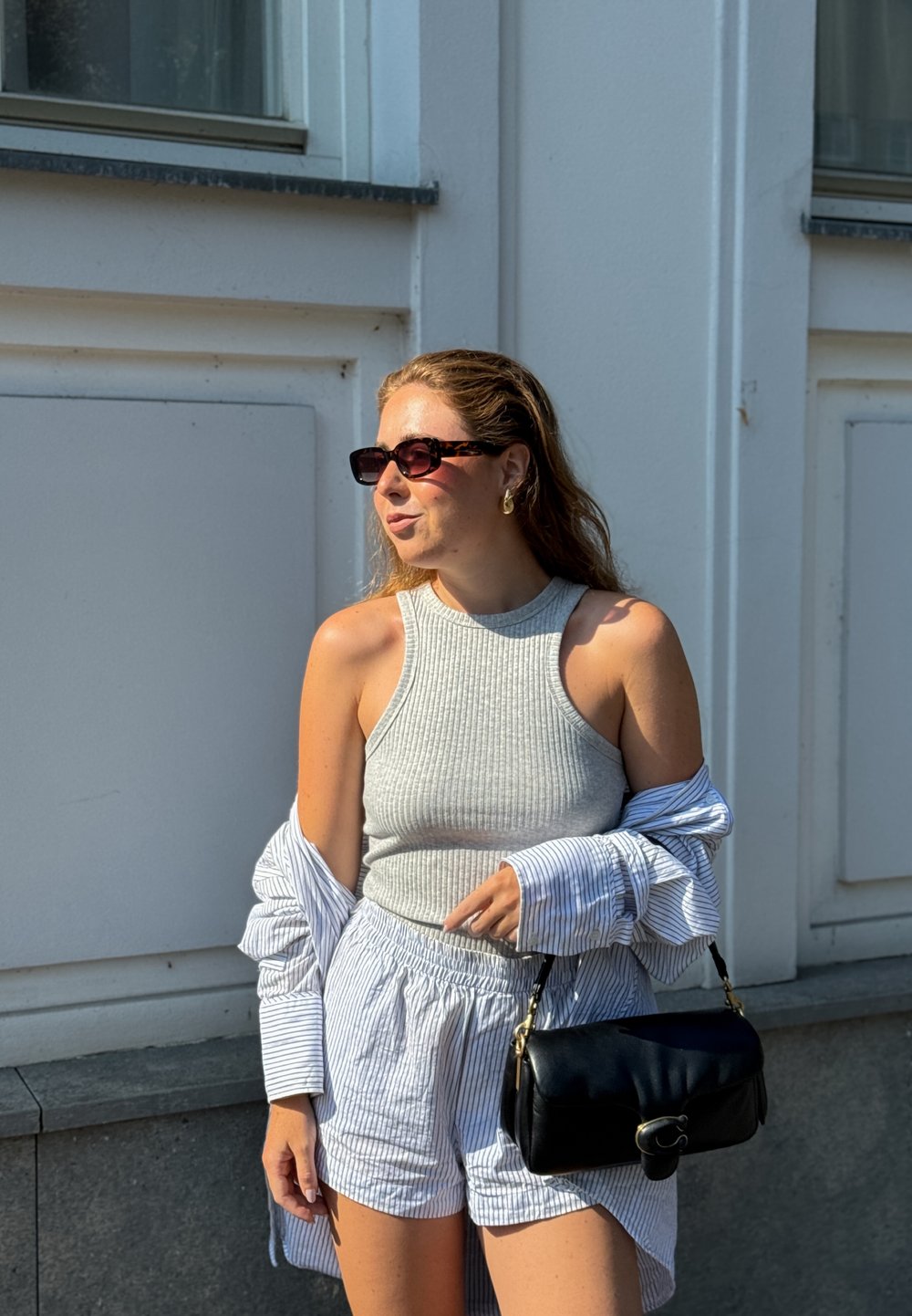 Grå rillede tanktop og stribede shorts, lagd med en oversized blå og hvid stribet skjorte. Sort skuldertaske og katteøje solbriller.