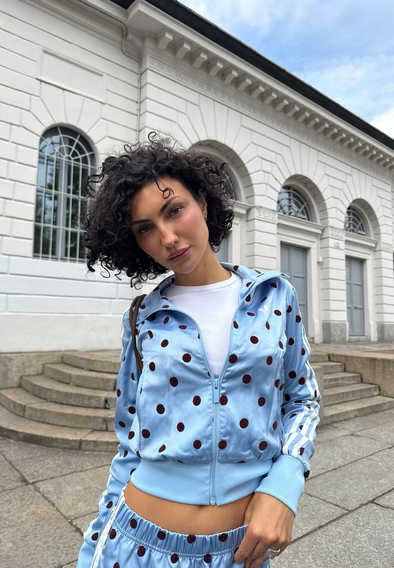 Femme aux cheveux bouclés portant un survêtement bleu à pois et une chemise blanche, debout devant un bâtiment blanc avec des fenêtres en arc et des marches.