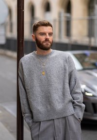 Pulover gri din tricot cu tiv și manșete cu nervuri, combinat cu pantaloni gri deschis. Accesoriile includ coliere aurii suprapuse și o suprafață texturată.