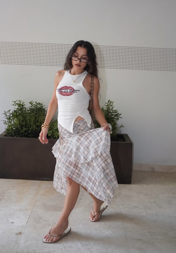 Mujer con gafas, top blanco sin mangas con estampado de labios, falda a cuadros y sandalias, está de pie en el interior sosteniendo su falda, cerca de plantas en macetas.