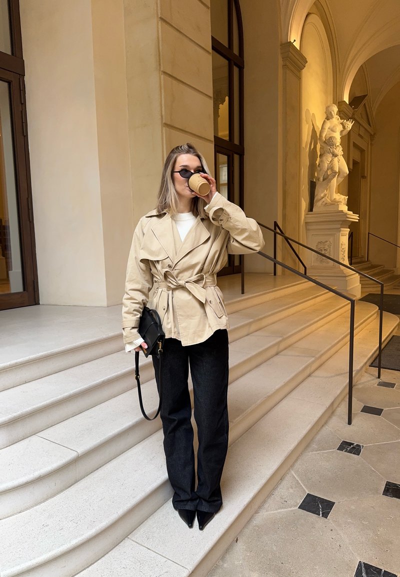 Femme en veste beige ceinturée et pantalon noir buvant dans une tasse, debout sur des marches en pierre à l'intérieur d'un bâtiment avec une statue et un arc en arrière-plan.